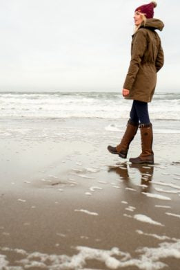 Woman wearing Ariat Argentium Parka in olive green walks along a winter beach, paired with boots and burgundy beanie
