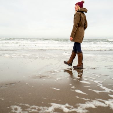 Woman wearing Ariat Argentium Parka in olive green walks along a winter beach, paired with boots and burgundy beanie