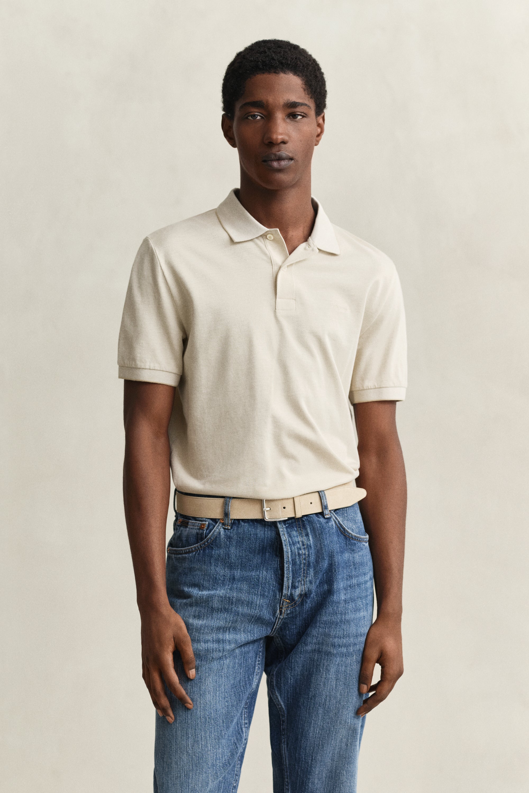 Man wearing GANT 2 Tone SS Polo in cream with collar, paired with blue jeans and beige belt against neutral background